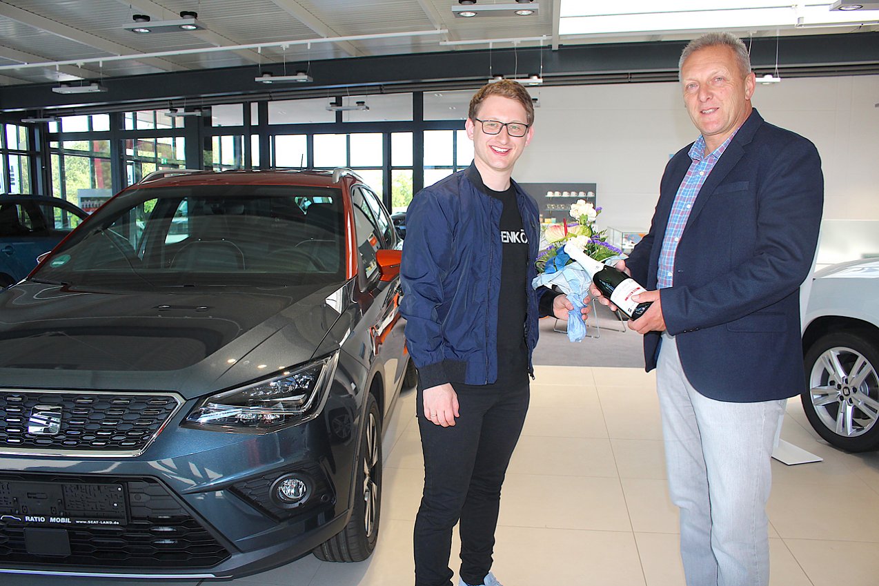 Der Preistr&auml;ger Samuel R&ouml;sch und Filialleiter von Annaberg R&uuml;diger Heinze stehen im Autohaus vor einem SEAT Modell. In der Hand halten Sie Blumen und eine Flasche. (Foto: RATIO MOBIL)